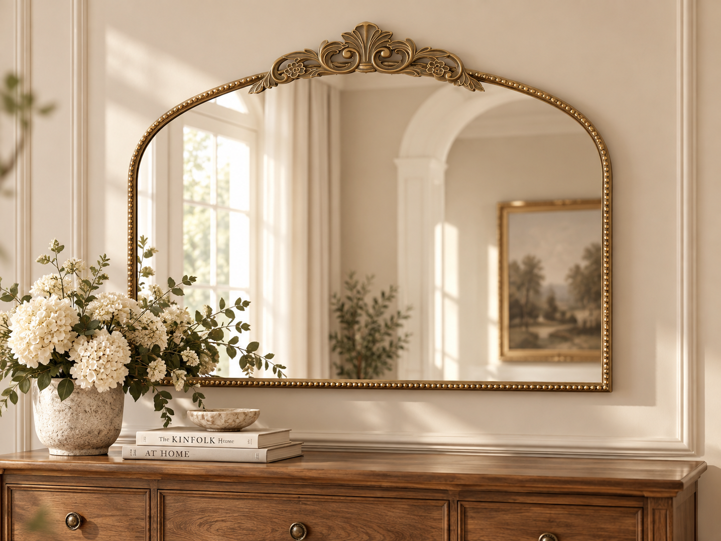 Gold ornate arched wall mirror styled above a console table in an elegant, light-filled room with soft floral accents.